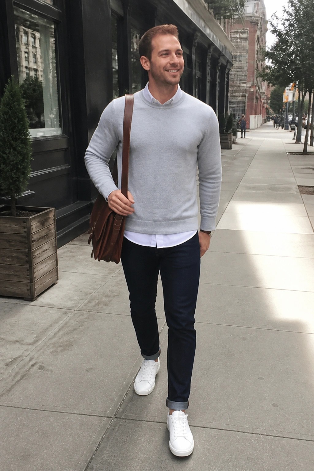 Man in light gray sweater and dark jeans carrying brown bag on city sidewalk