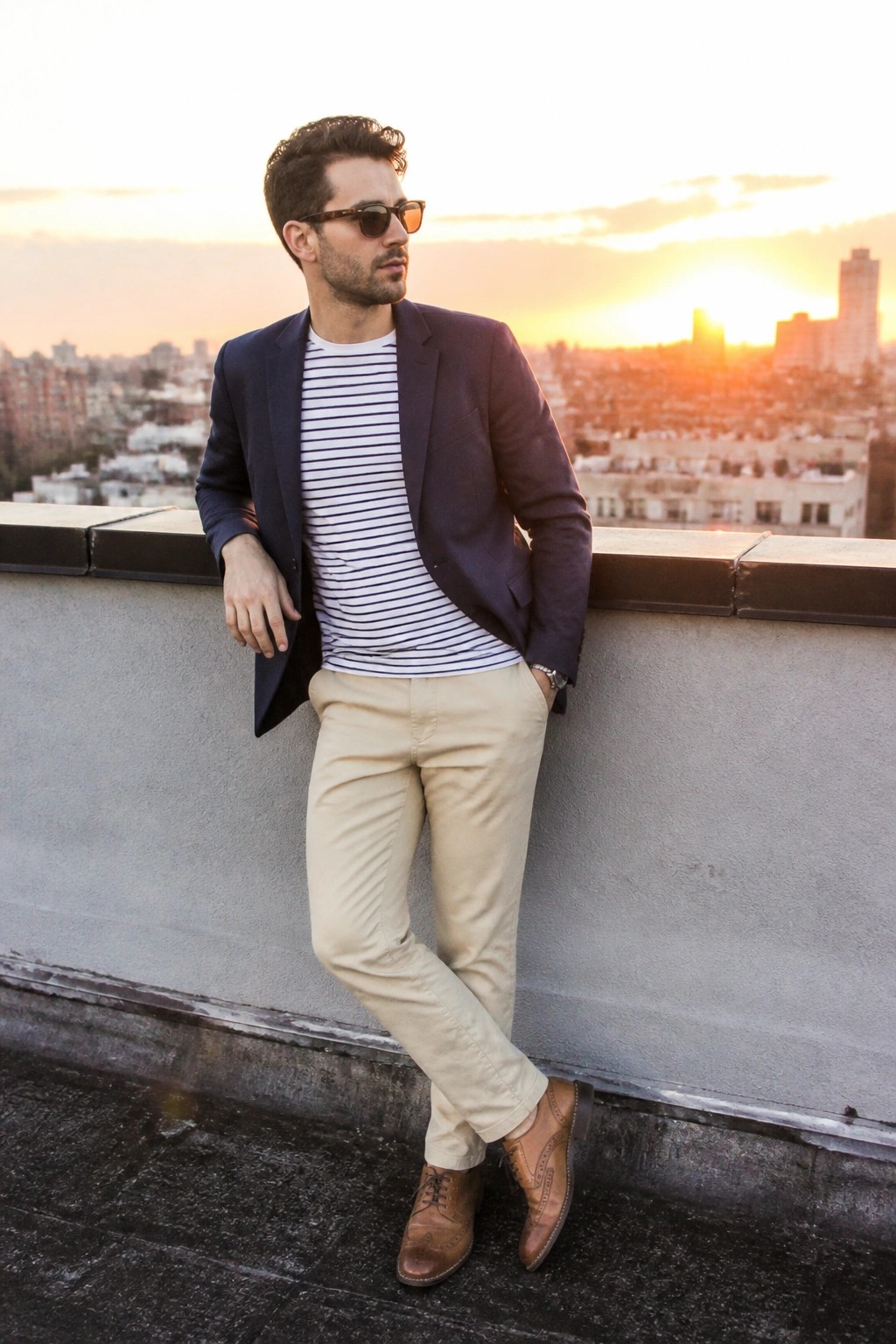 Man in navy blazer, striped shirt, and beige pants on rooftop at sunset