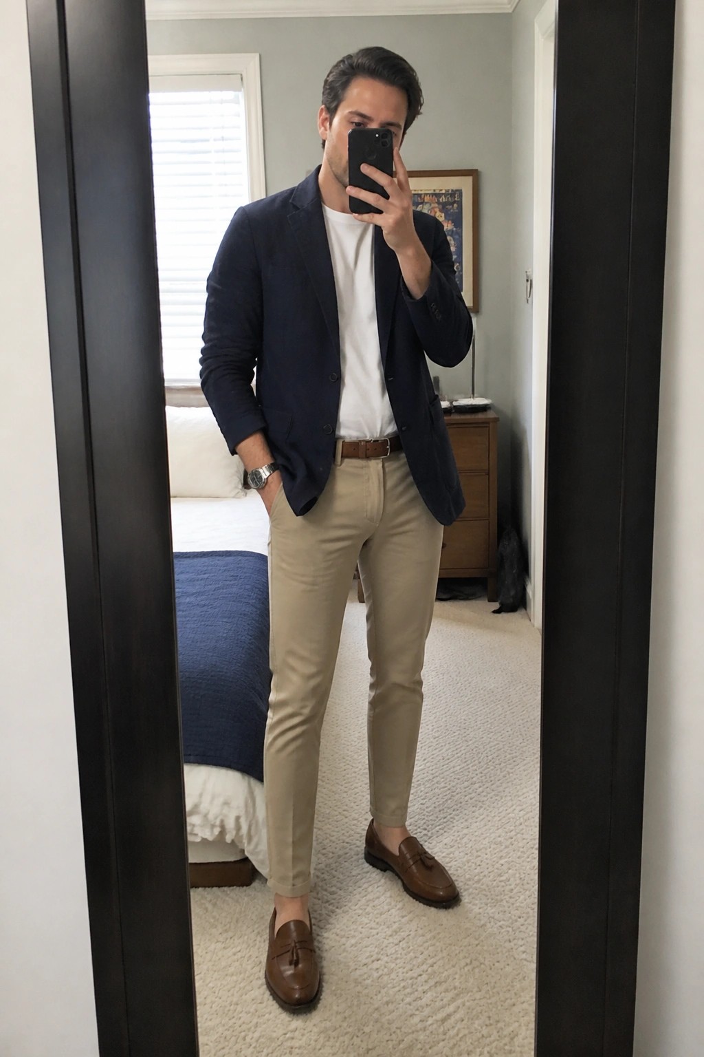 Man in navy blazer and khaki pants taking mirror selfie in bedroom