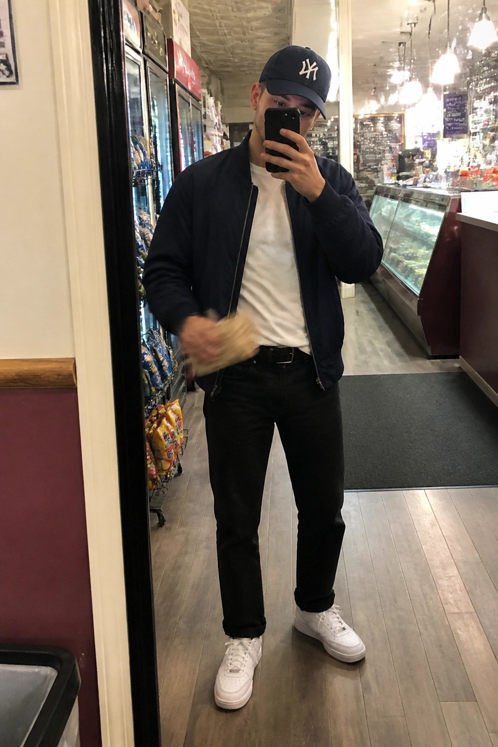 Man in navy bomber jacket over white t-shirt with black jeans, white sneakers, and navy cap taking a mirror selfie in a store.