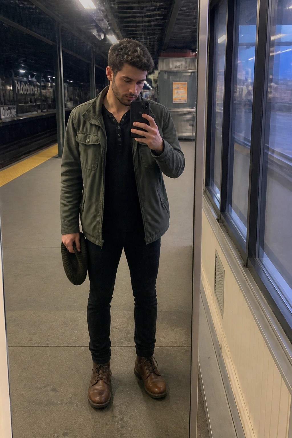 Man in green jacket takes mirror selfie on train platform holding beanie