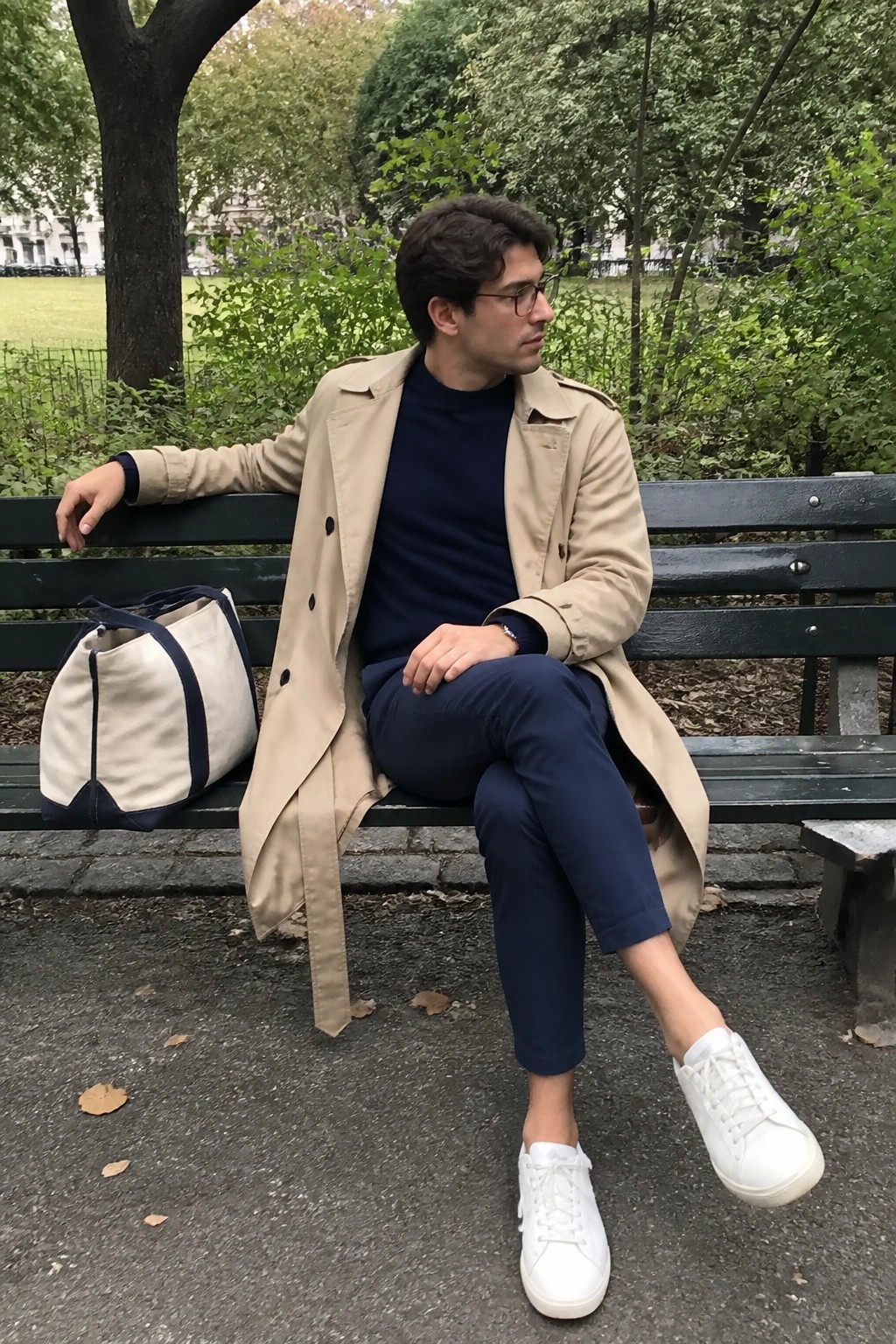 Man in beige trench coat on green park bench beside canvas tote bag