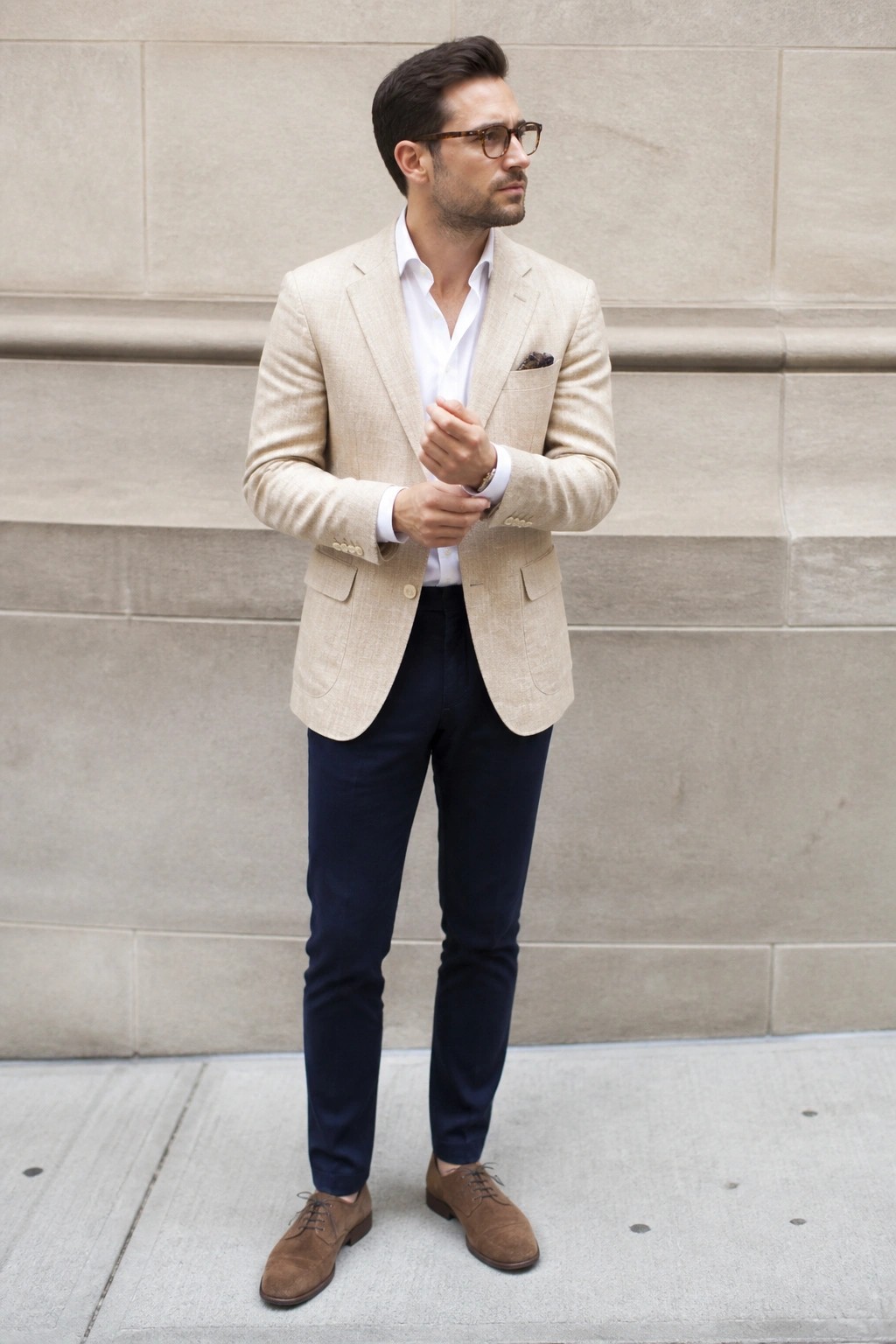 Man in light beige blazer white shirt navy pants and brown suede shoes standing against stone wall with hands clasped