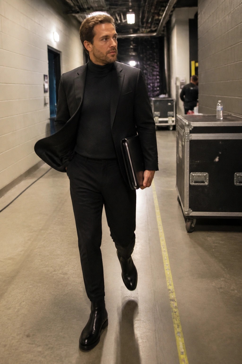 Man in black suit and turtleneck walking through industrial hallway with folder