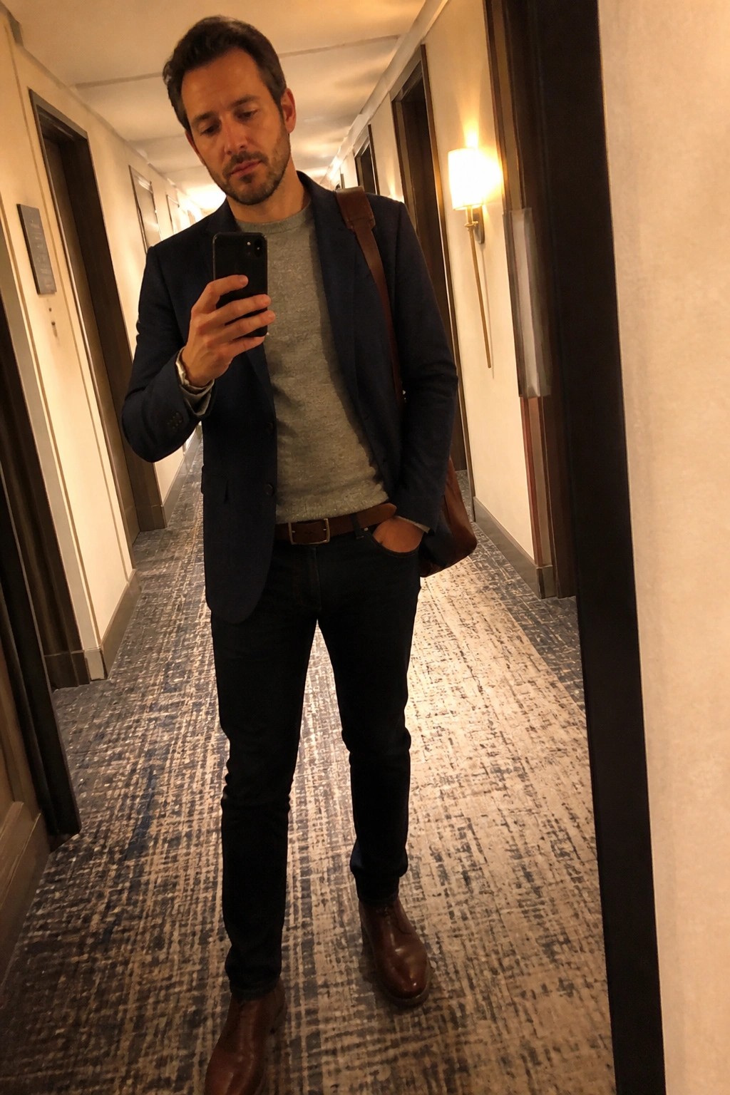 Man in navy blazer and jeans taking mirror selfie in hallway
