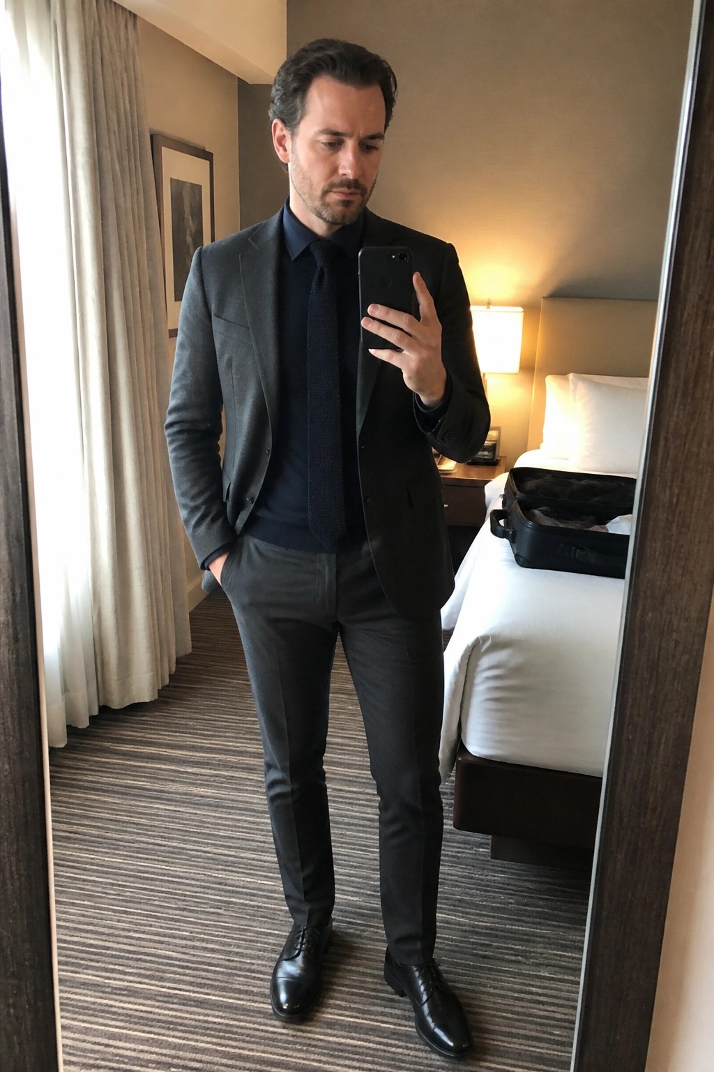 A man wearing a charcoal gray suit over a navy sweater and matching tie with black dress shoes taking a mirror selfie in a hotel room.