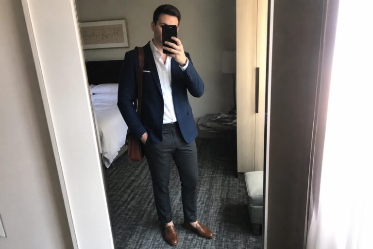 Man in navy blazer, white dress shirt, gray trousers, brown belt, brown leather loafers, and brown leather bag taking a mirror selfie in a hotel room.
