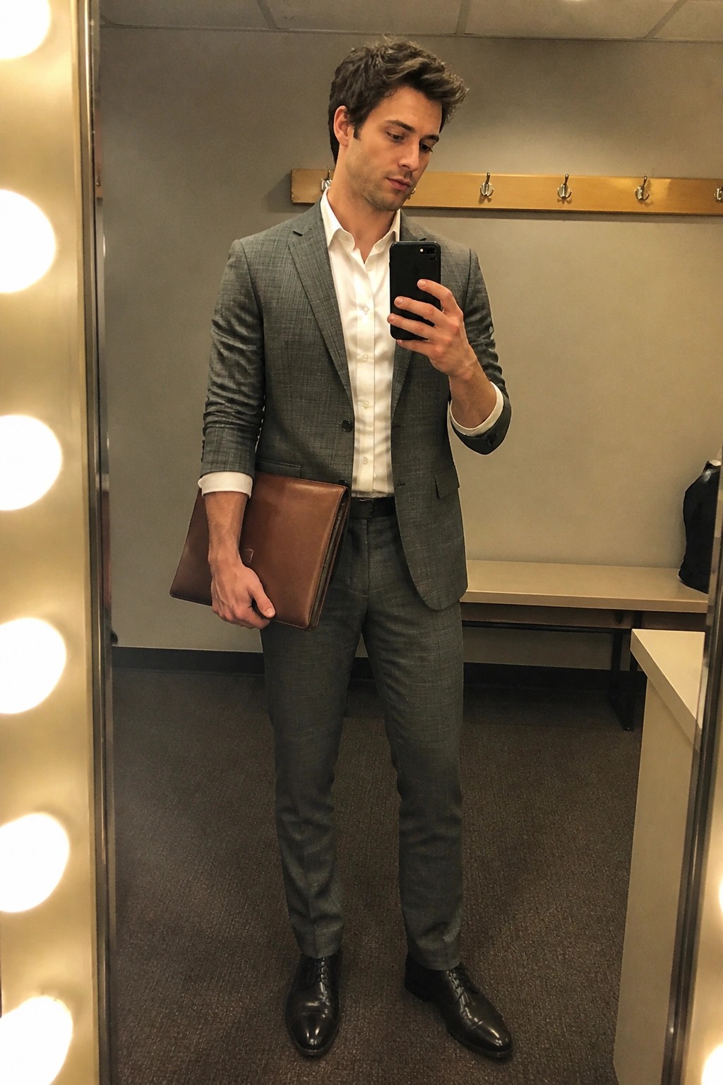 Man in gray plaid suit taking mirror selfie while holding brown leather portfolio