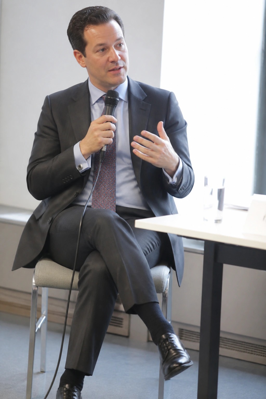 Man in dark suit holds microphone and gestures while seated at table