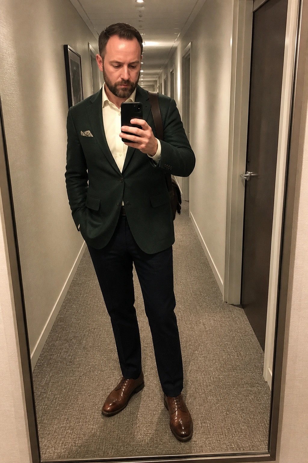 Man in green blazer and navy pants taking mirror selfie in hallway