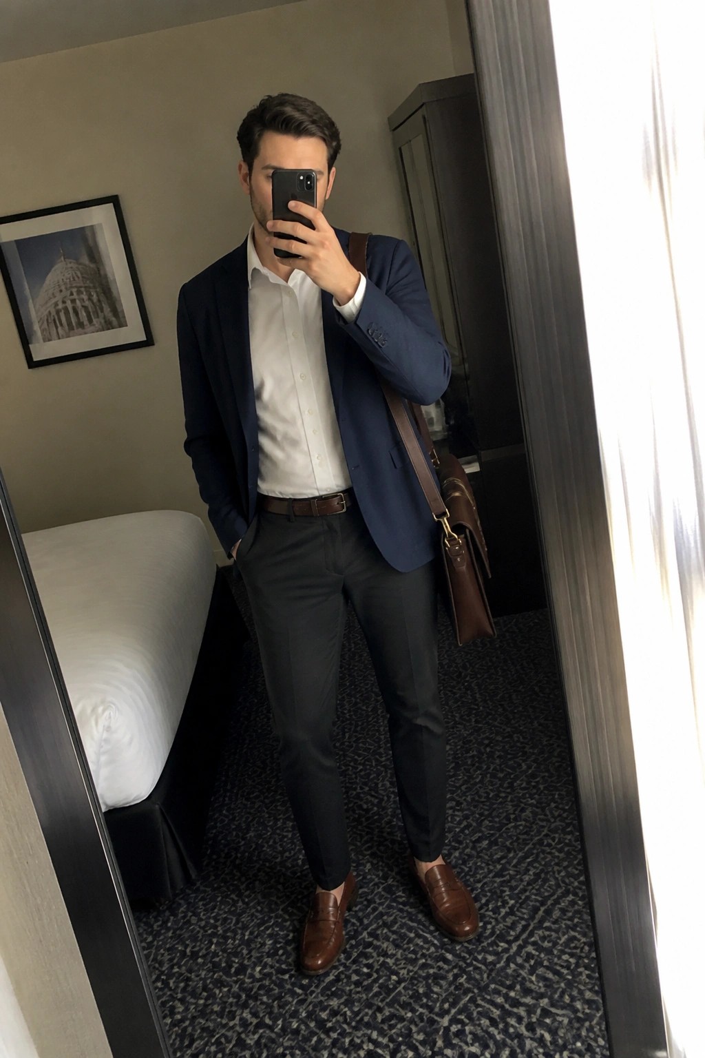 Man in navy blazer, white dress shirt, gray trousers, brown belt, brown leather loafers, and brown leather bag taking a mirror selfie in a hotel room.