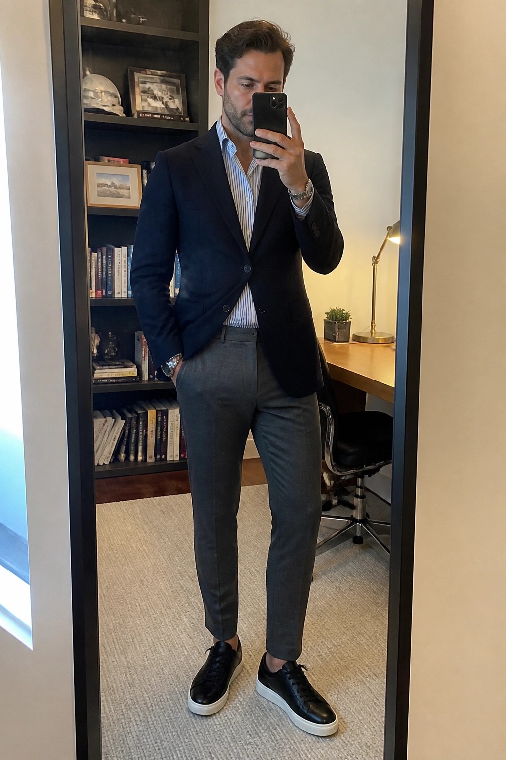 Man in navy blazer, striped shirt, gray trousers, and black sneakers takes mirror selfie