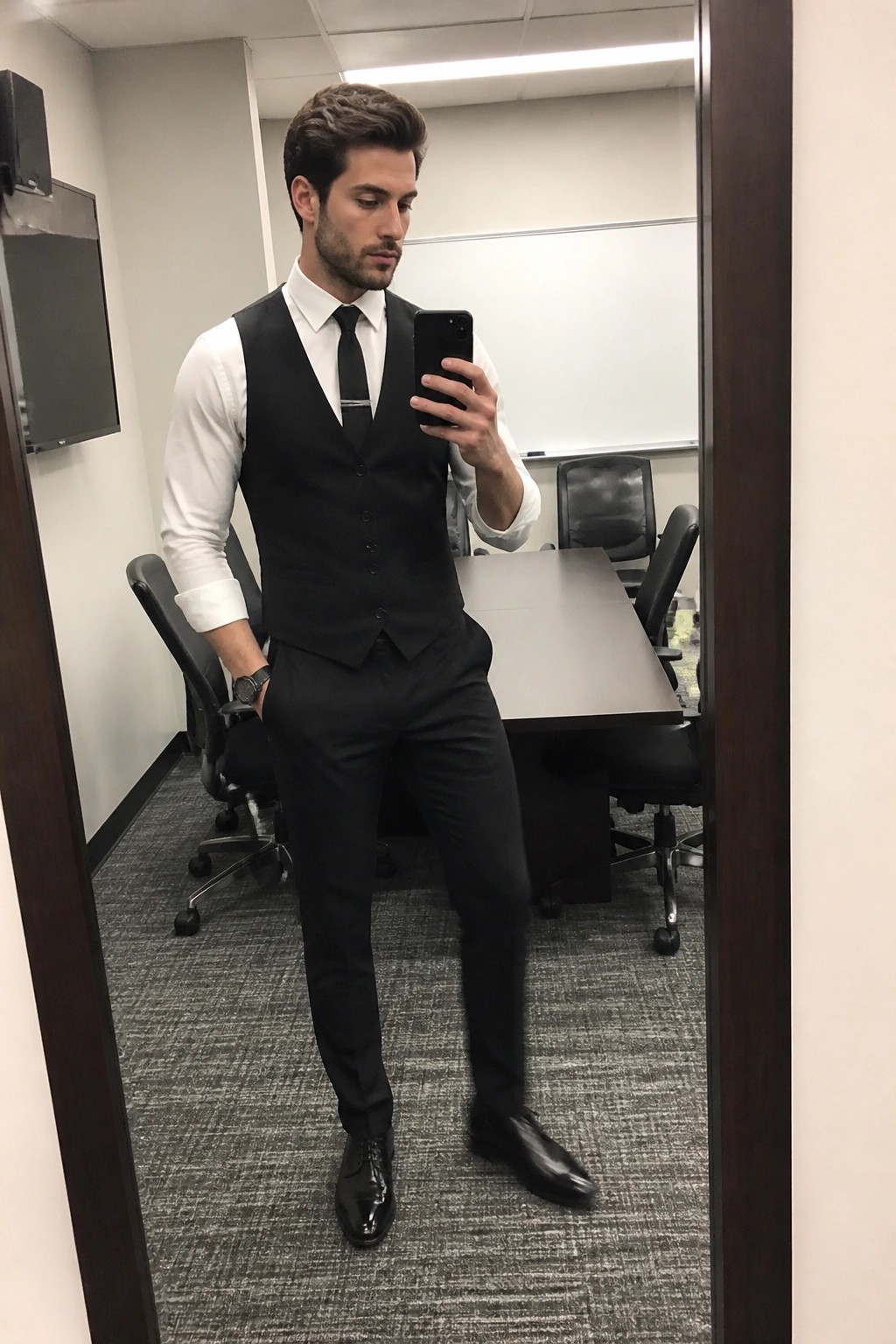 Man in white shirt, black vest, and tie taking mirror selfie in conference room