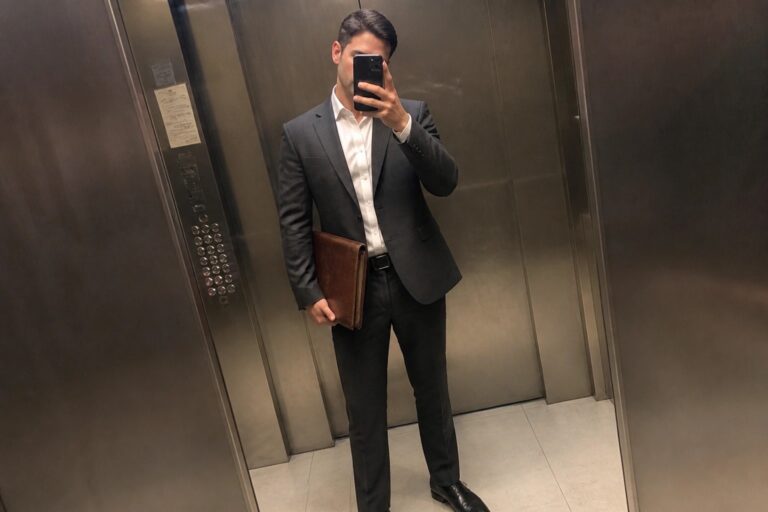 22 Polished Business Professional Outfits for Men in Modern Corporate Offices Man in a dark gray business suit jacket and trousers with a white button-down shirt black dress shoes and brown leather portfolio bag taking a mirror selfie in an elevator.