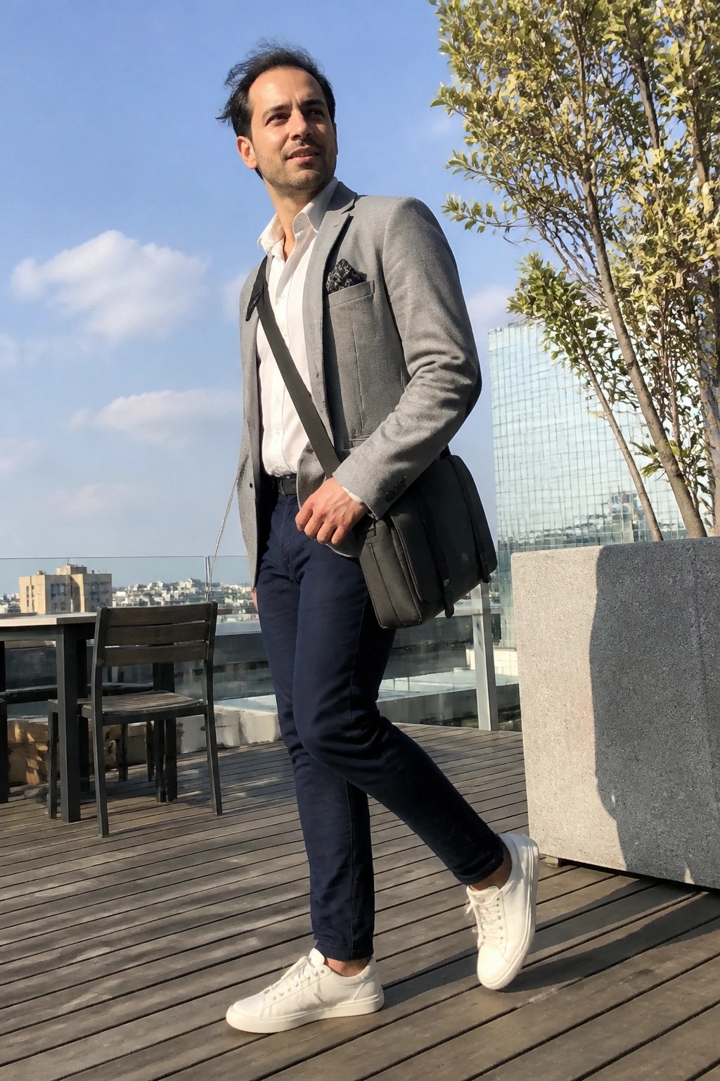 Man in gray blazer, blue pants, white sneakers with black bag on sunny rooftop