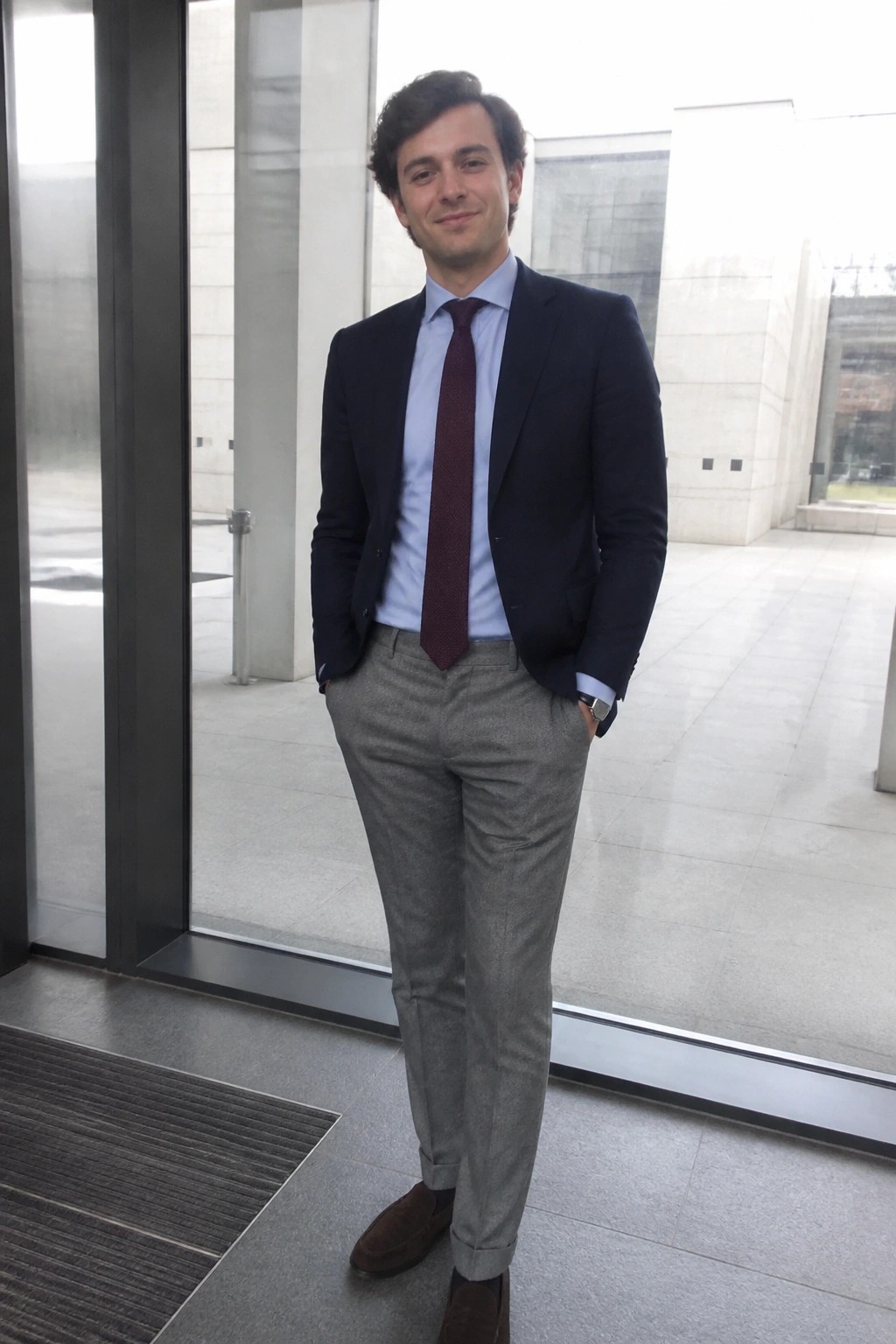 Man wearing navy blazer over light blue shirt with maroon tie, gray trousers, and brown suede shoes in indoor office setting.