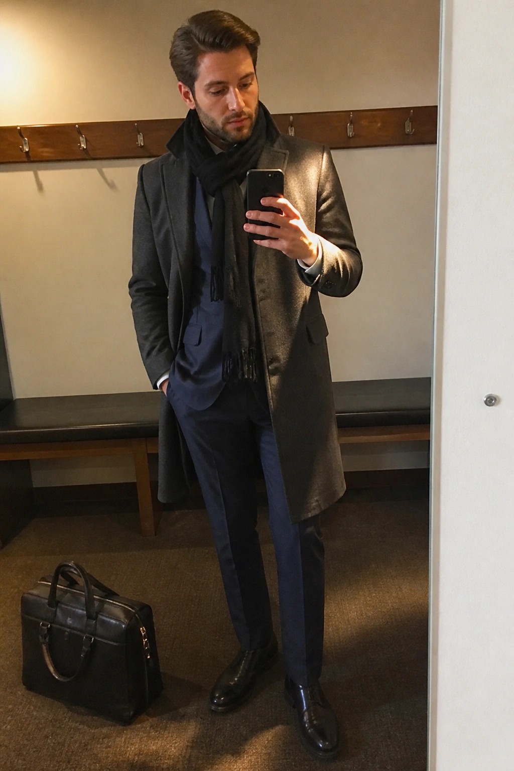 Man in gray overcoat and blue suit with black scarf taking mirror selfie near briefcase