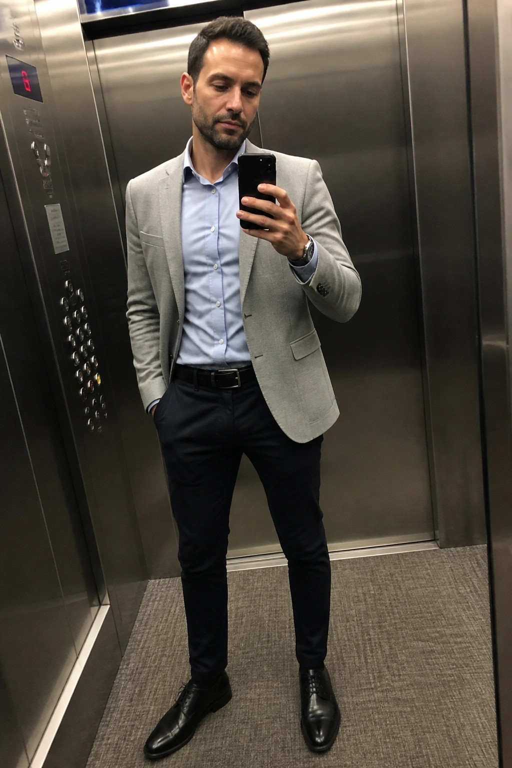 Man standing in elevator wearing light gray blazer over light blue button down shirt with navy trousers black belt and black dress shoes while holding phone for mirror selfie.