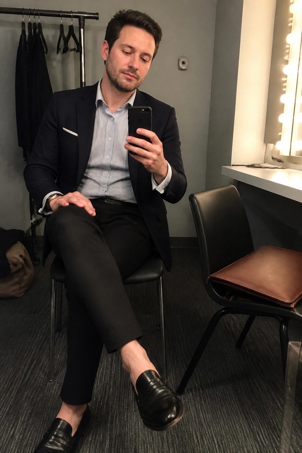 Man in navy blazer sits holding smartphone in room with clothing rack and vanity mirror