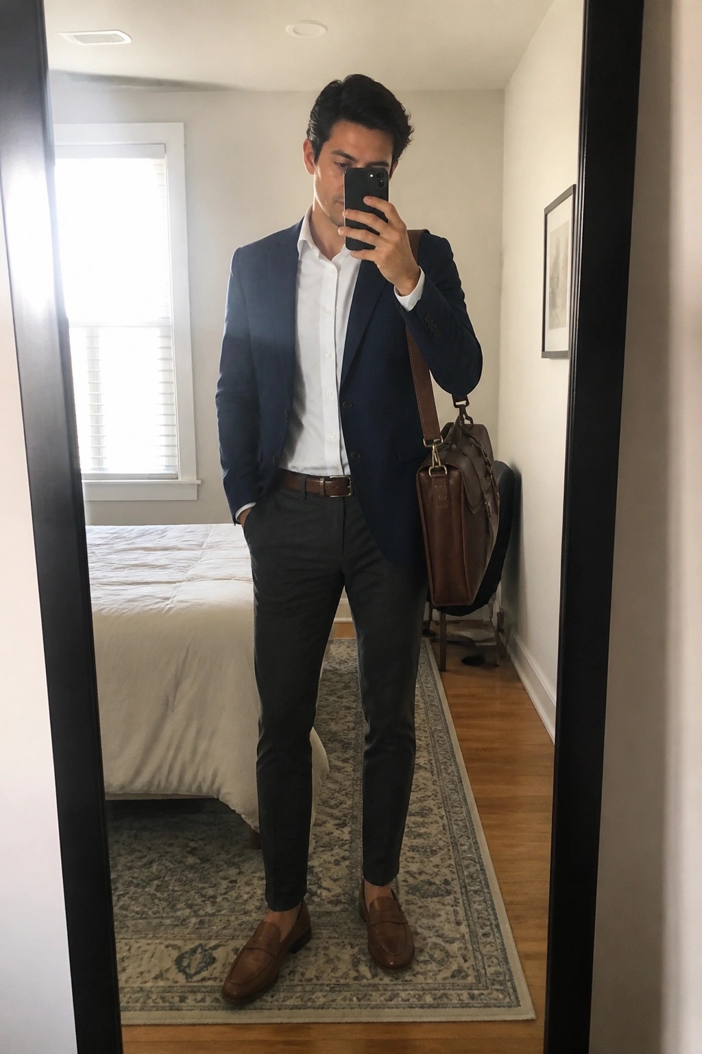 Man in navy blazer and gray pants taking mirror selfie with brown leather bag.
