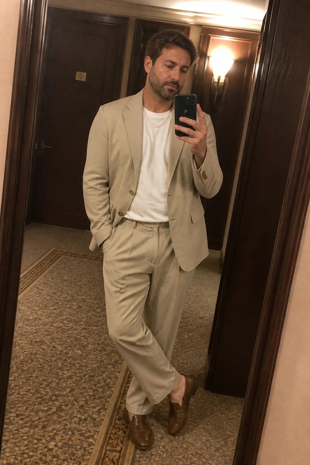 Man in beige suit and white tee taking mirror selfie in hallway