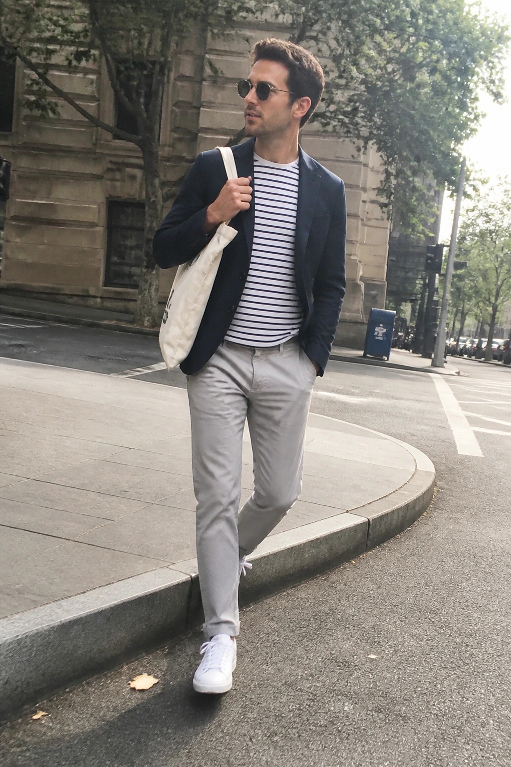 Man in navy blazer over striped tee with gray pants and tote bag on city street