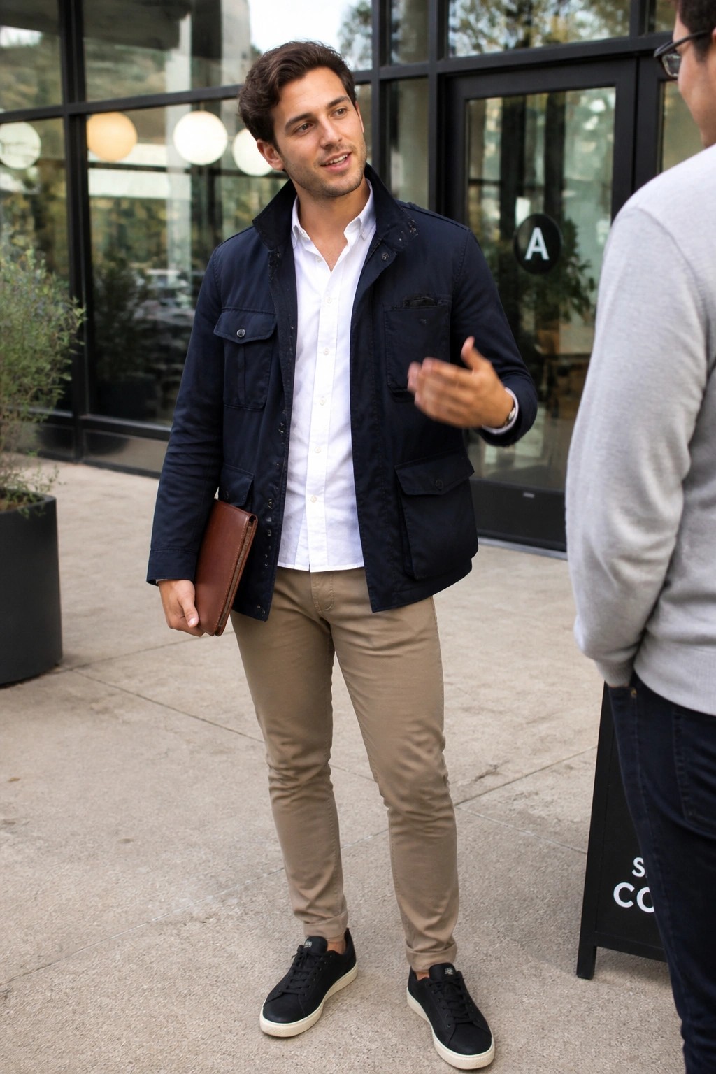 Man in navy utility jacket over white button down shirt, khaki pants, and black sneakers holding brown leather folder outdoors near building entrance.