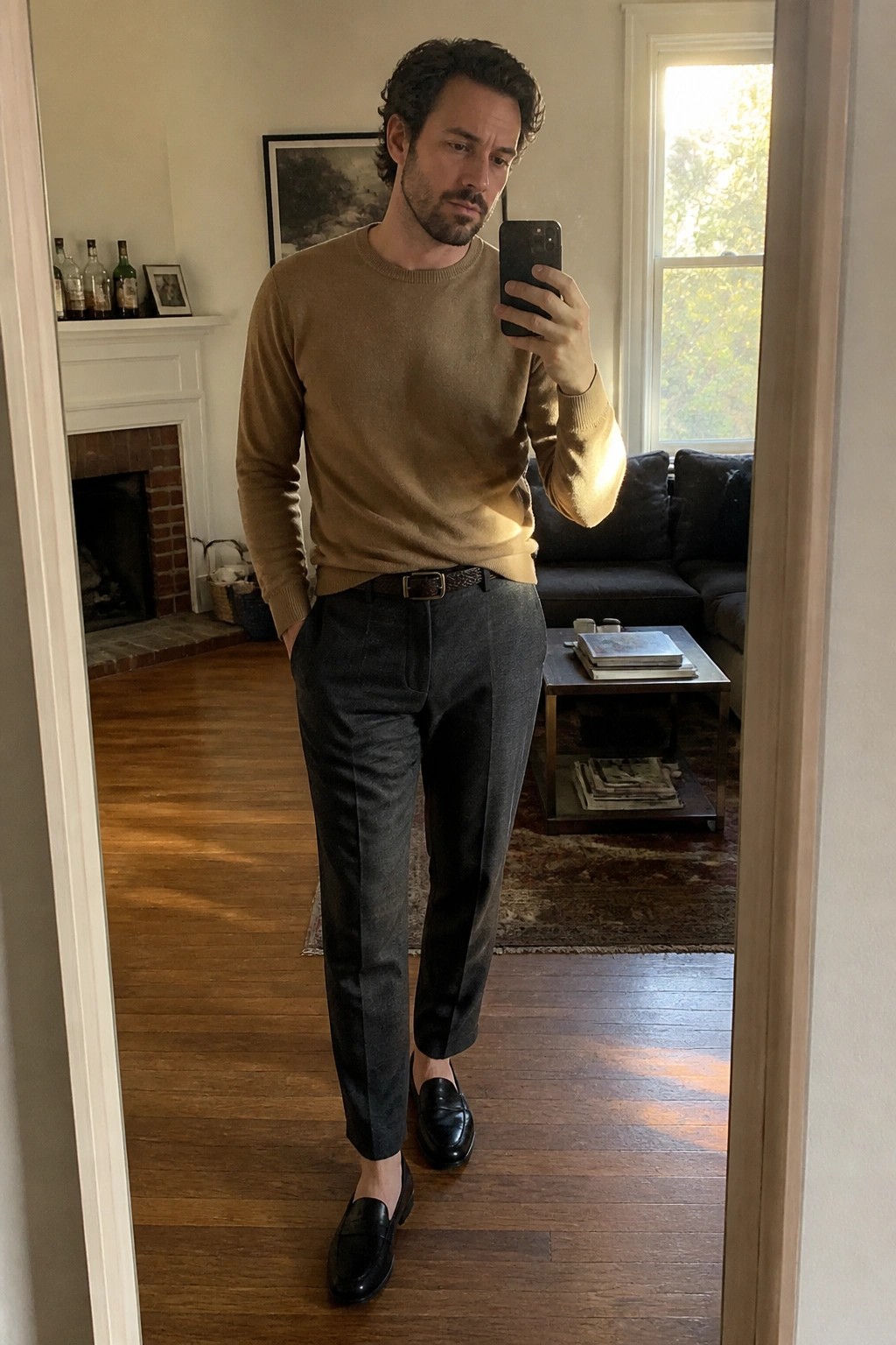 Man in tan sweater and gray pants taking mirror selfie in living room