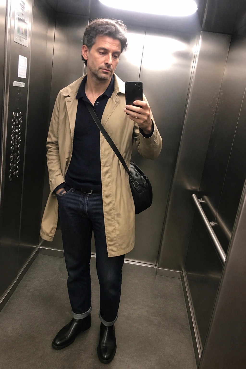 Man in beige trench coat taking phone selfie in metal elevator with black bag