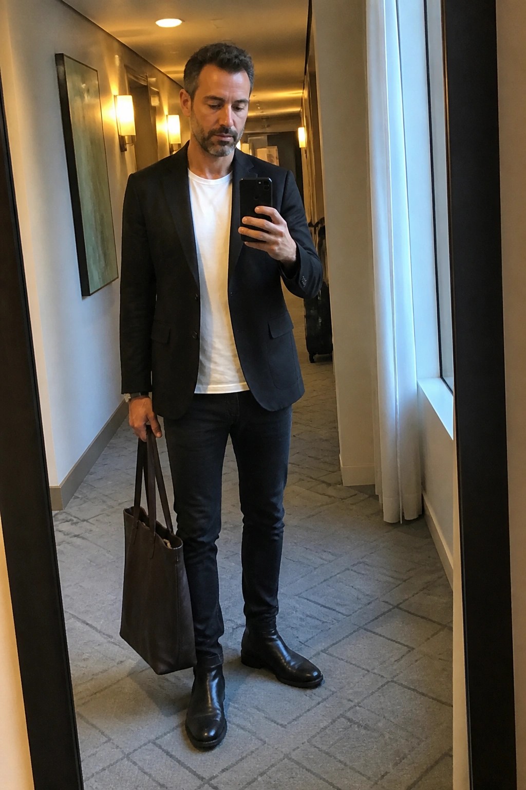 Man in black blazer and jeans takes mirror selfie holding brown leather tote.