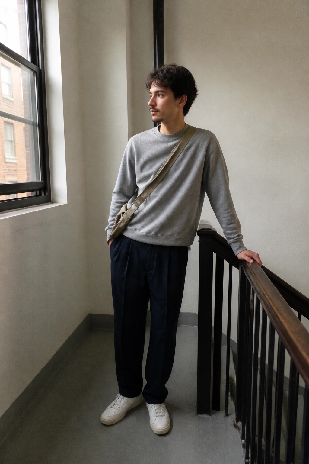 Man in gray sweatshirt with crossbody bag leaning on stair railing in navy pants