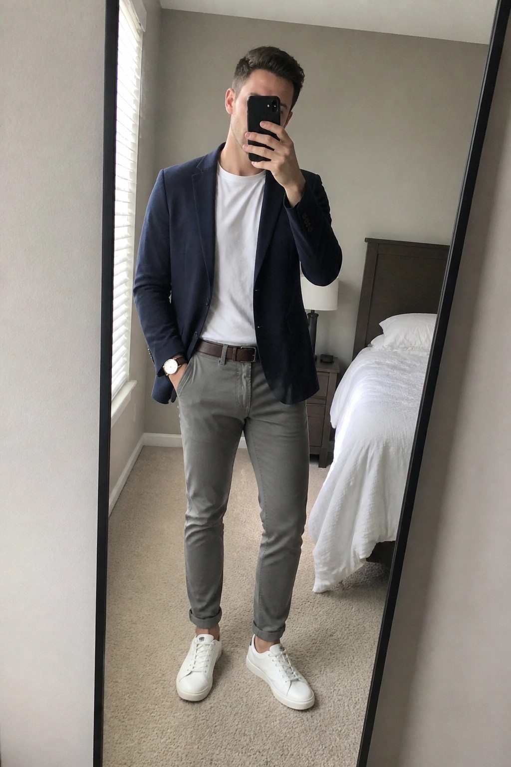 Man in navy blazer, white tee, and gray pants taking mirror selfie in bedroom