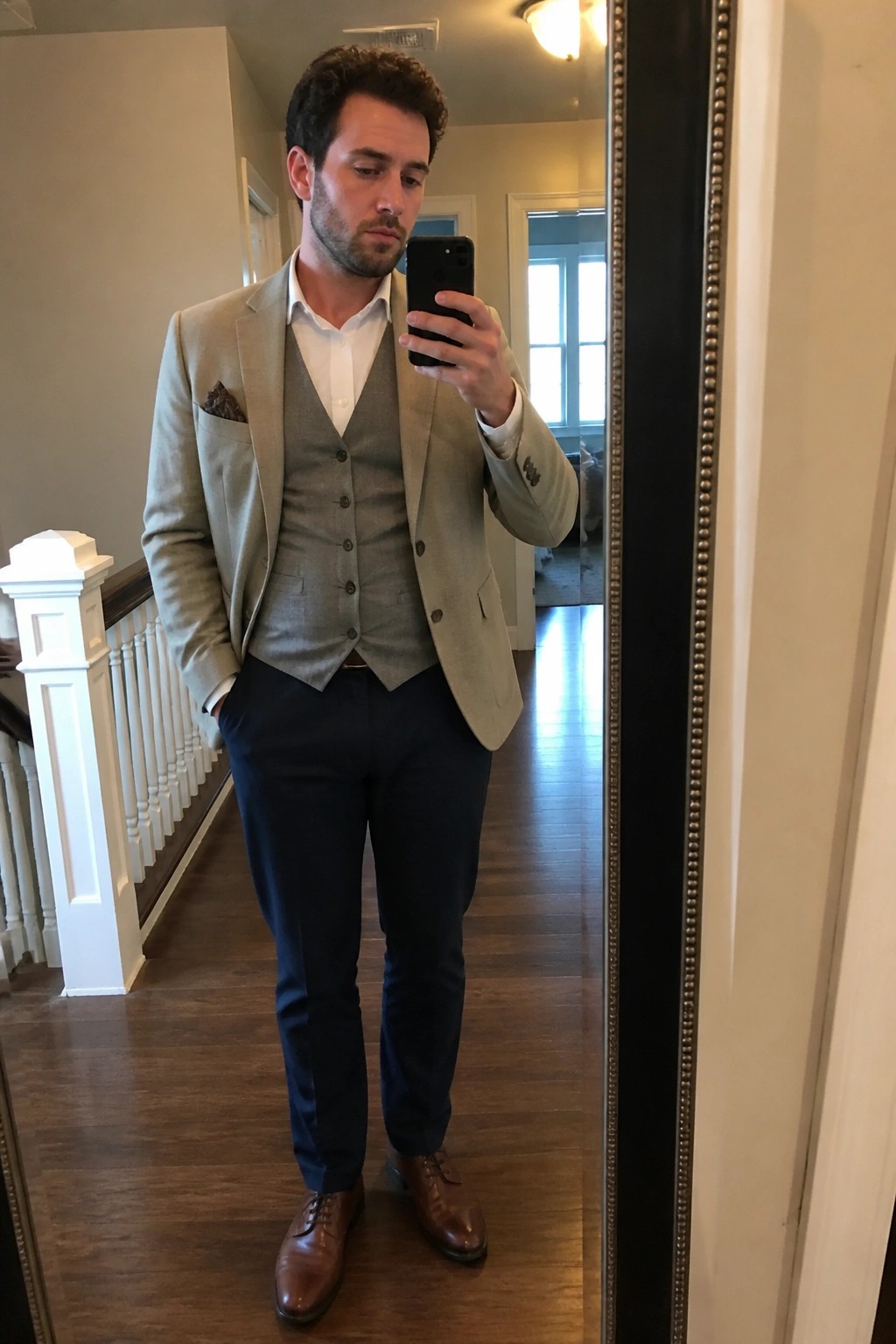 A man in a light beige blazer layered over a gray vest and white shirt paired with navy trousers and brown leather shoes taking a mirror selfie.