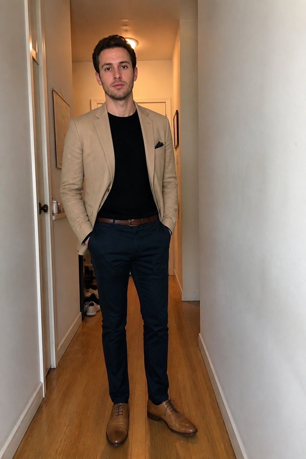 Man in beige blazer, black shirt, navy pants, and brown shoes in hallway.