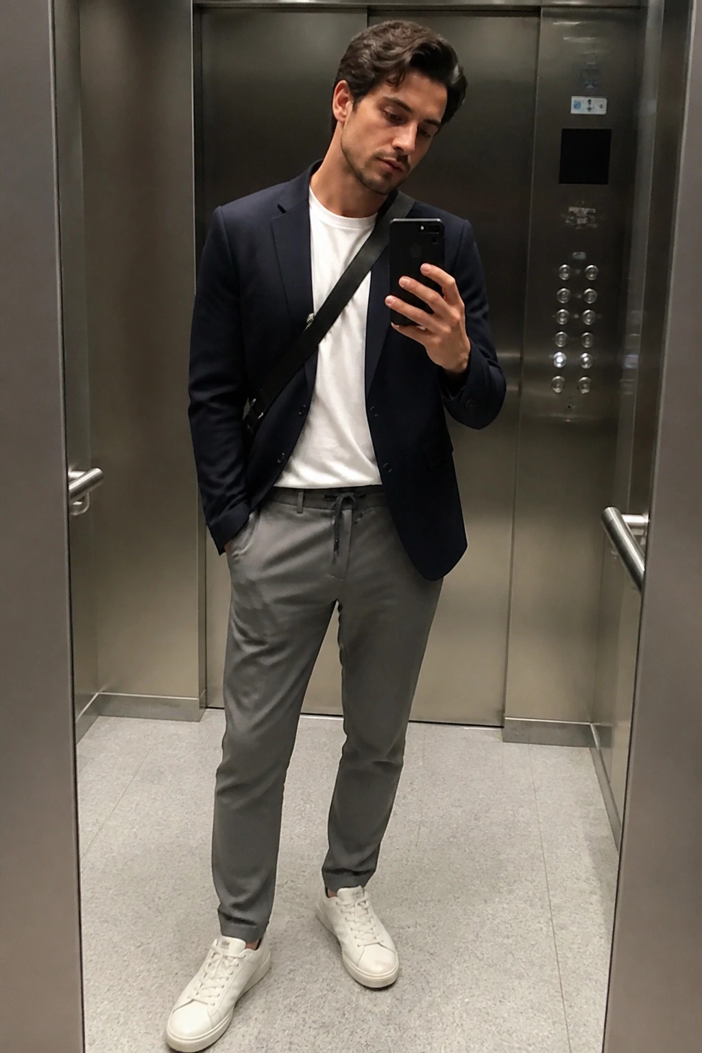 Man in navy blazer, white t-shirt, gray pants taking phone selfie in elevator