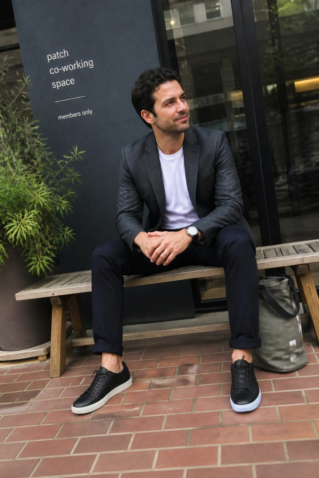 Man in gray blazer and black sneakers sits on bench with green bag