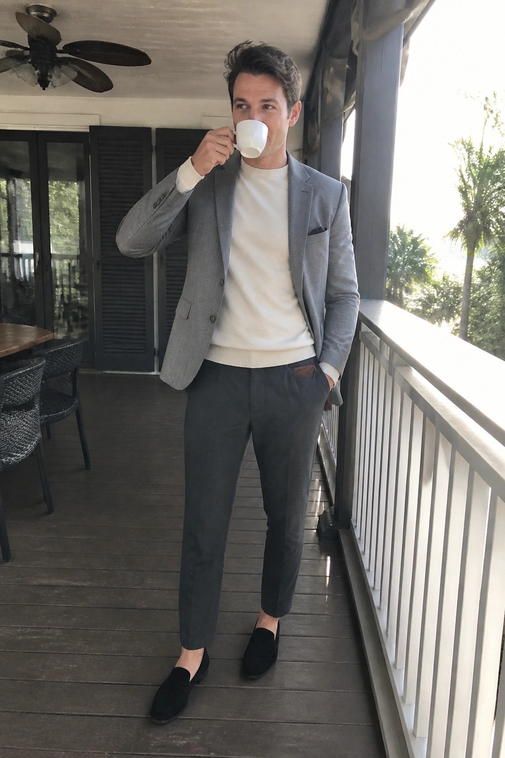 Man in gray blazer over white sweater sipping coffee on wooden porch