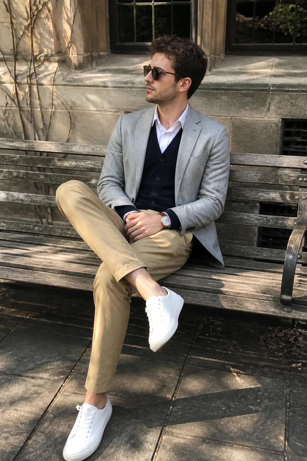 Man in light gray blazer, navy vest, khaki pants, and white sneakers on bench
