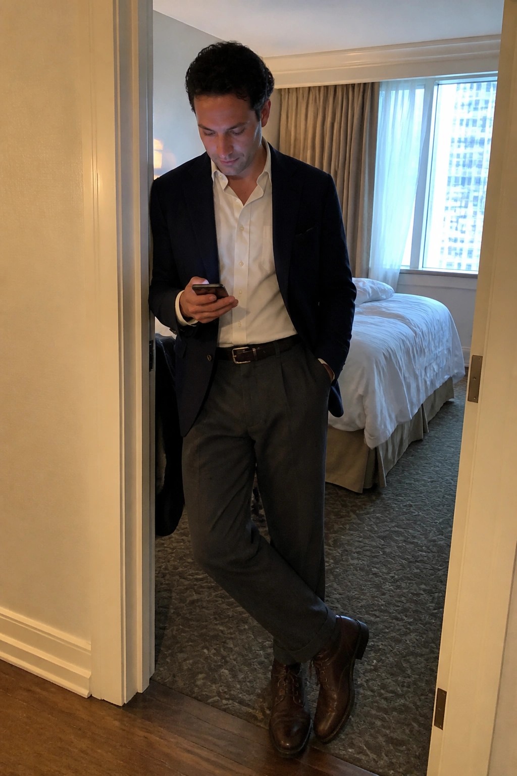 Man in navy blazer and gray pants checks phone in hotel room doorway