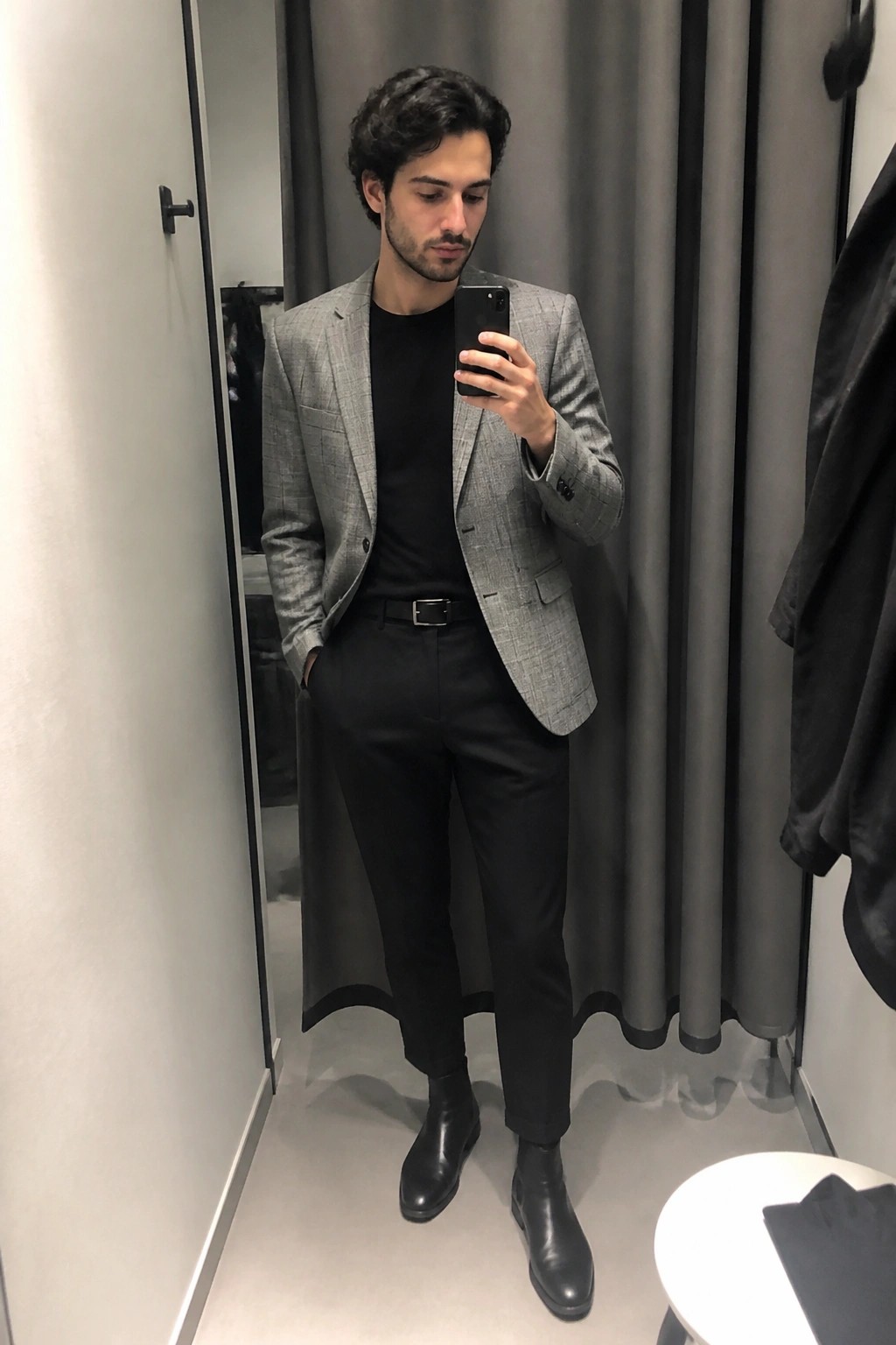 Man in gray plaid blazer, black shirt and pants taking mirror selfie in fitting room