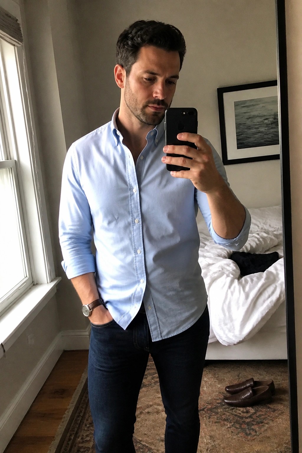 Man in light blue button-down shirt taking mirror selfie in bedroom with iPhone