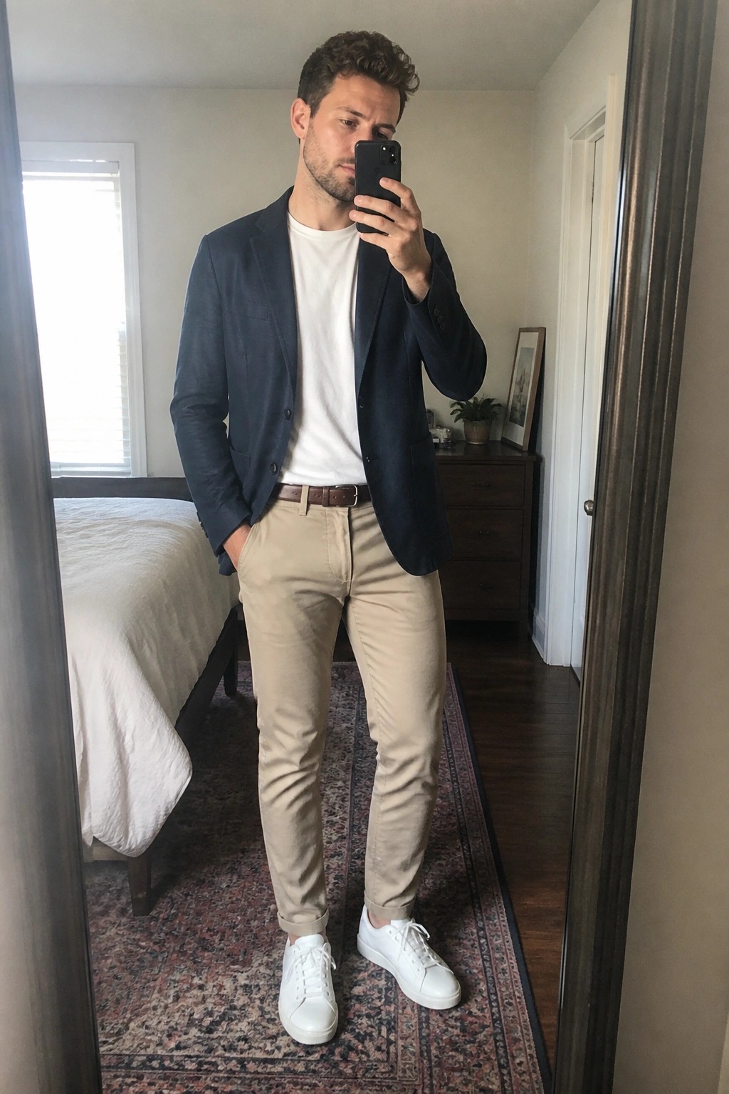 Man in navy blazer, white tee, khaki pants, and white sneakers taking mirror selfie