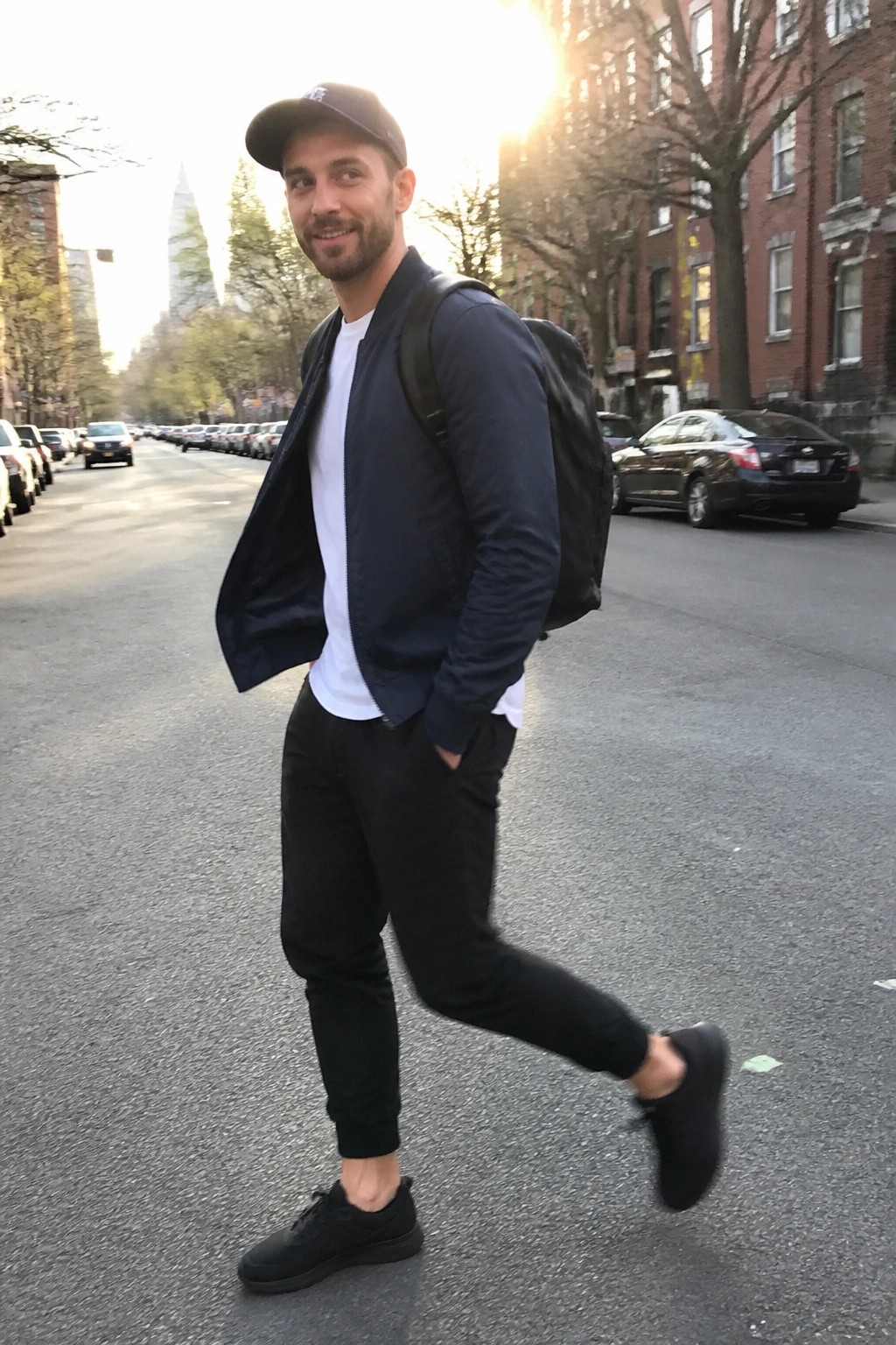 Smiling man in navy bomber jacket, cap, and backpack posing on sunny city street.
