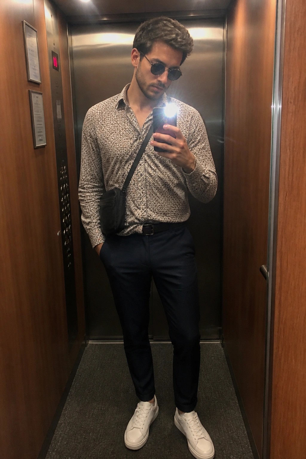 Man in patterned shirt, sunglasses, and white sneakers taking selfie in elevator