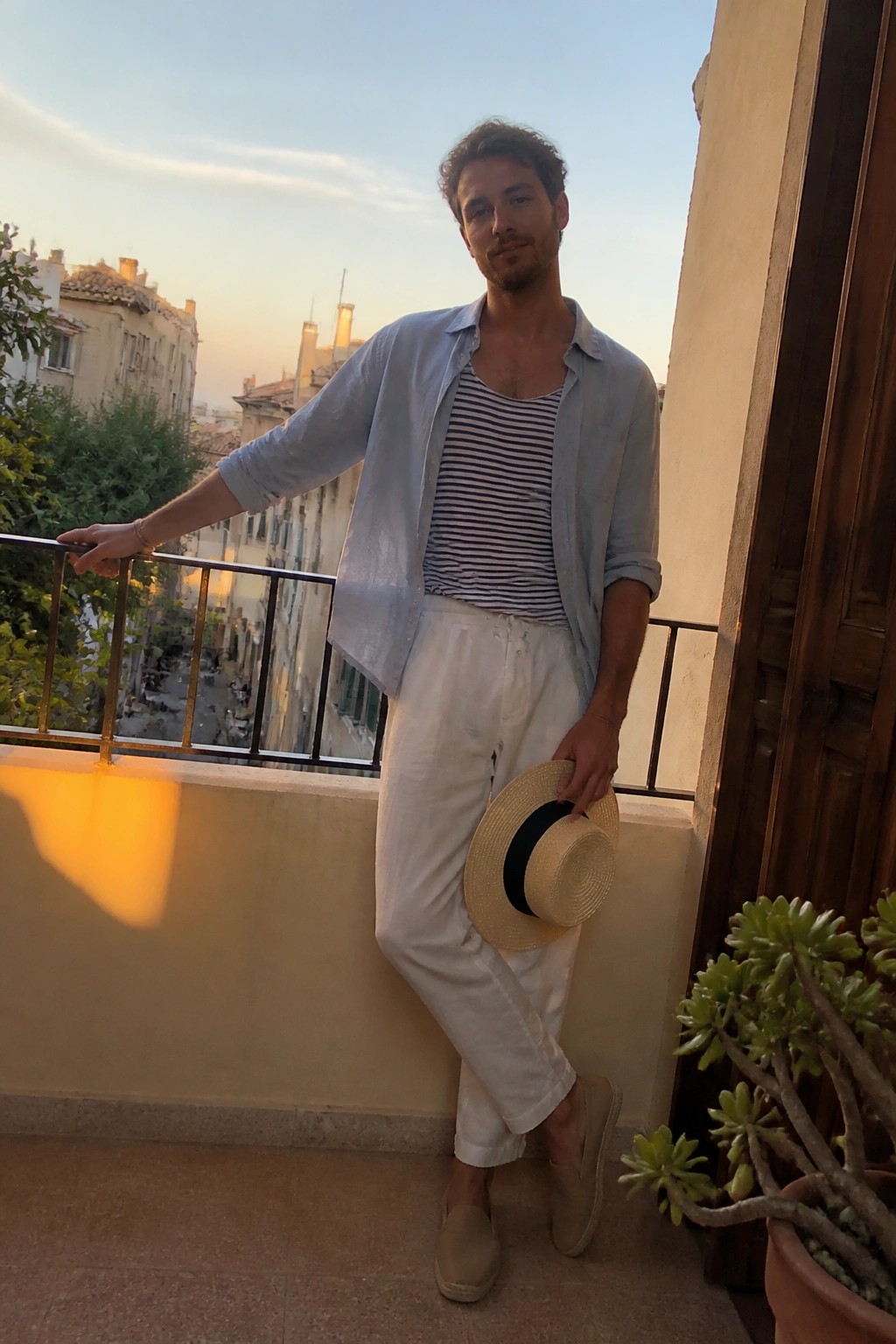 Man standing on a balcony wearing an open light blue shirt over a navy striped tank top, paired with white pants, beige espadrilles, and holding a straw hat.