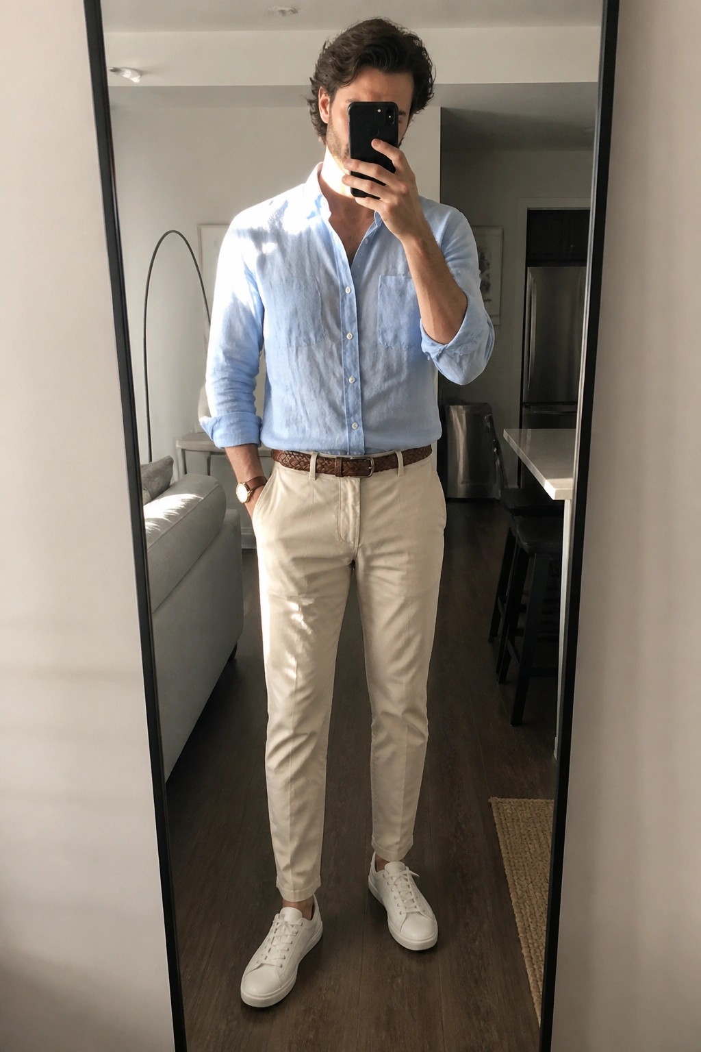 Man in light blue button down shirt tucked into cream pants with brown belt and white sneakers taking mirror selfie