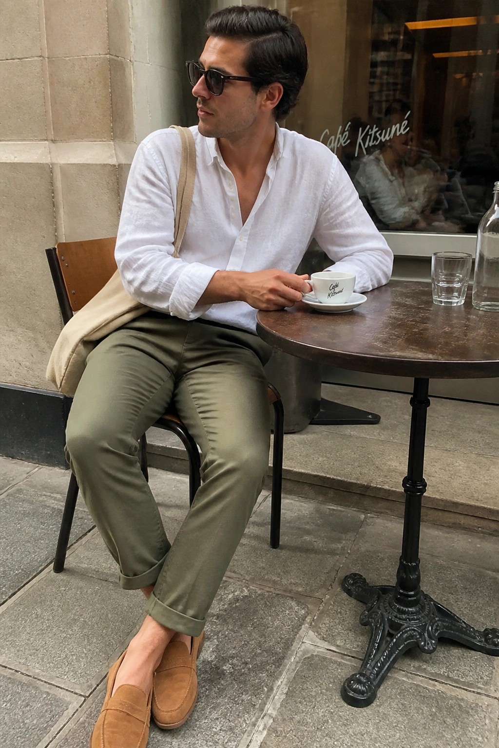 Man in white shirt and olive pants holds coffee at outdoor café table.