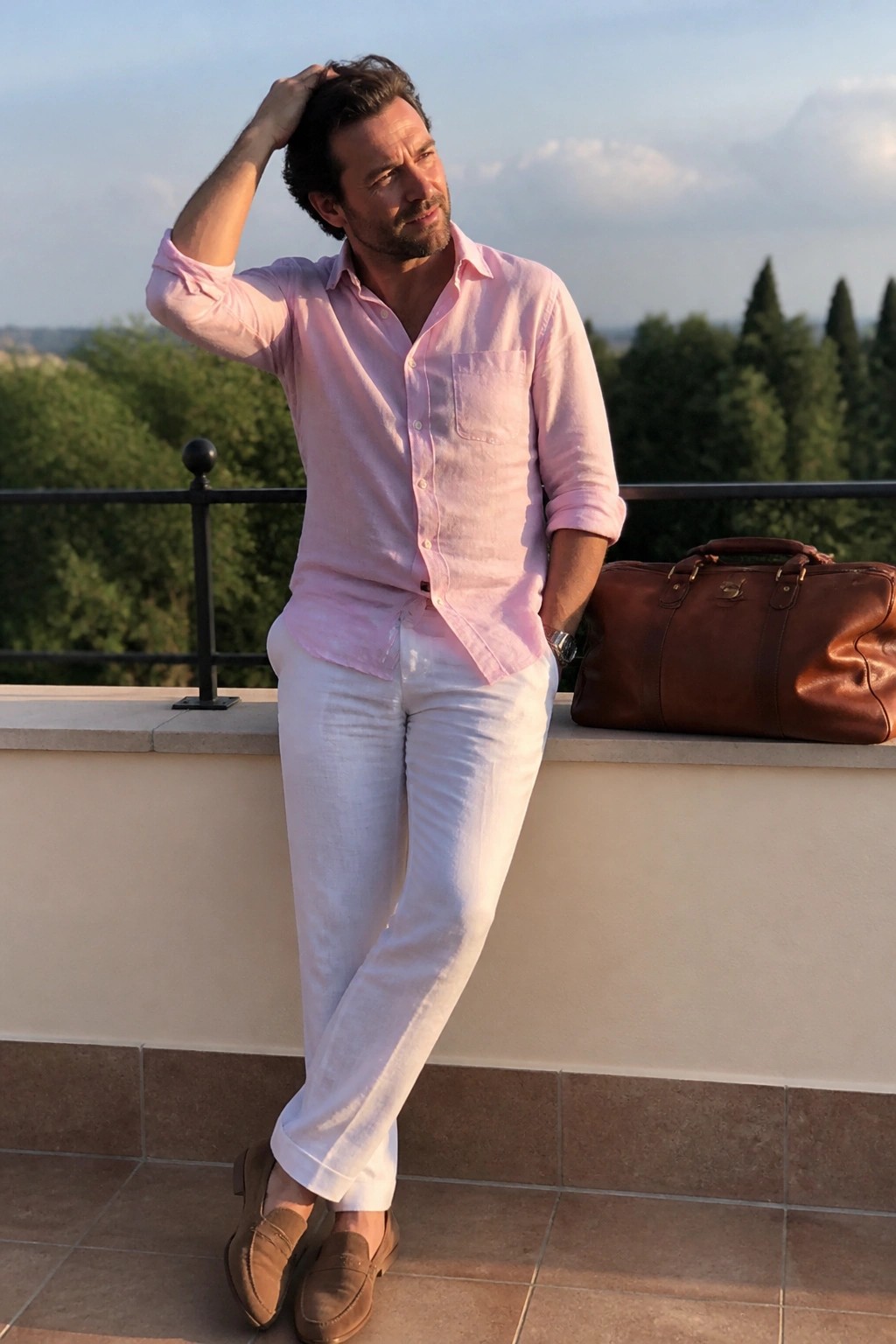 Man wearing light pink linen button down shirt with rolled sleeves, white linen trousers, brown suede loafers and carrying a brown leather duffel bag in a smart casual summer outfit.