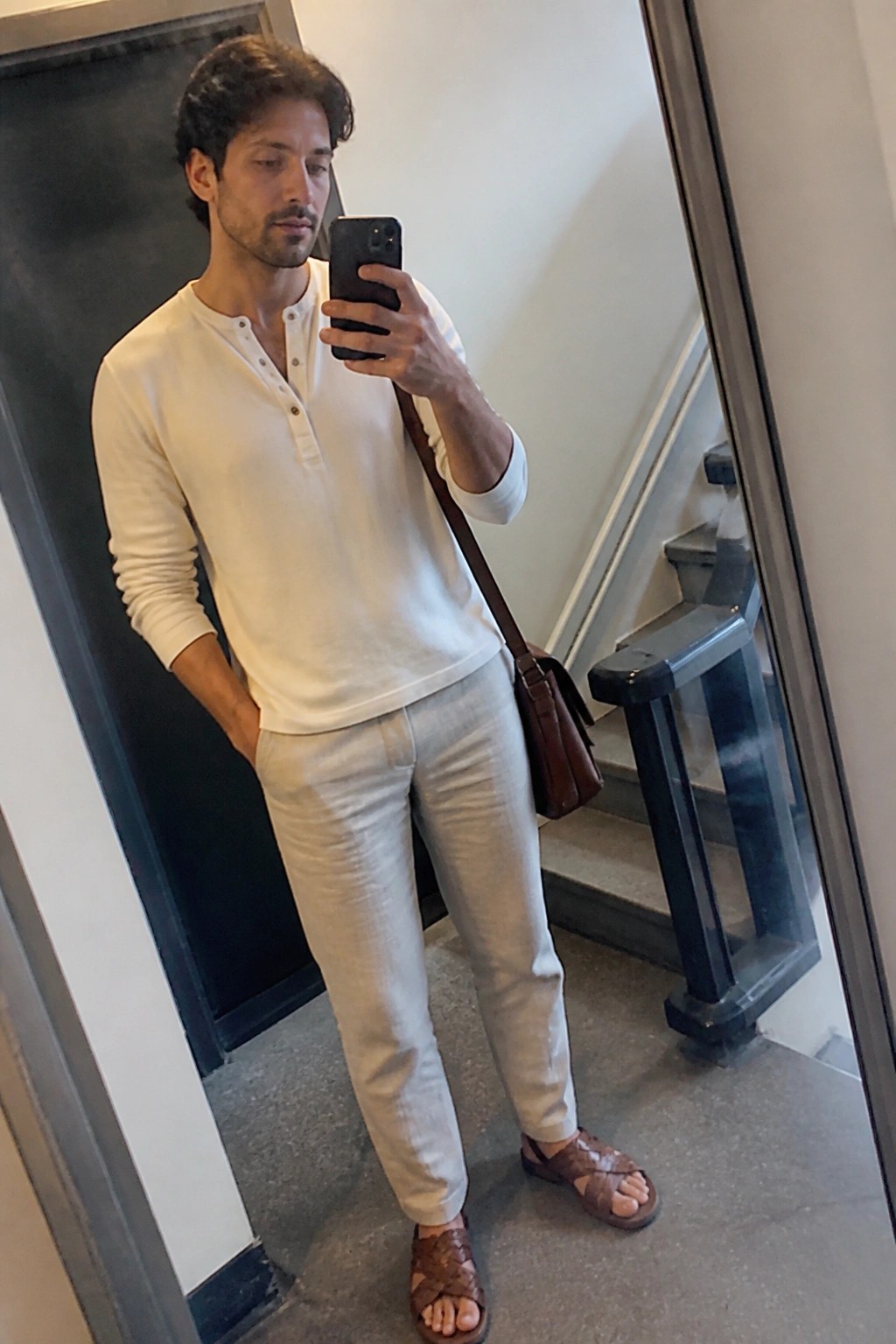Man in white henley shirt and light pants taking mirror selfie with phone