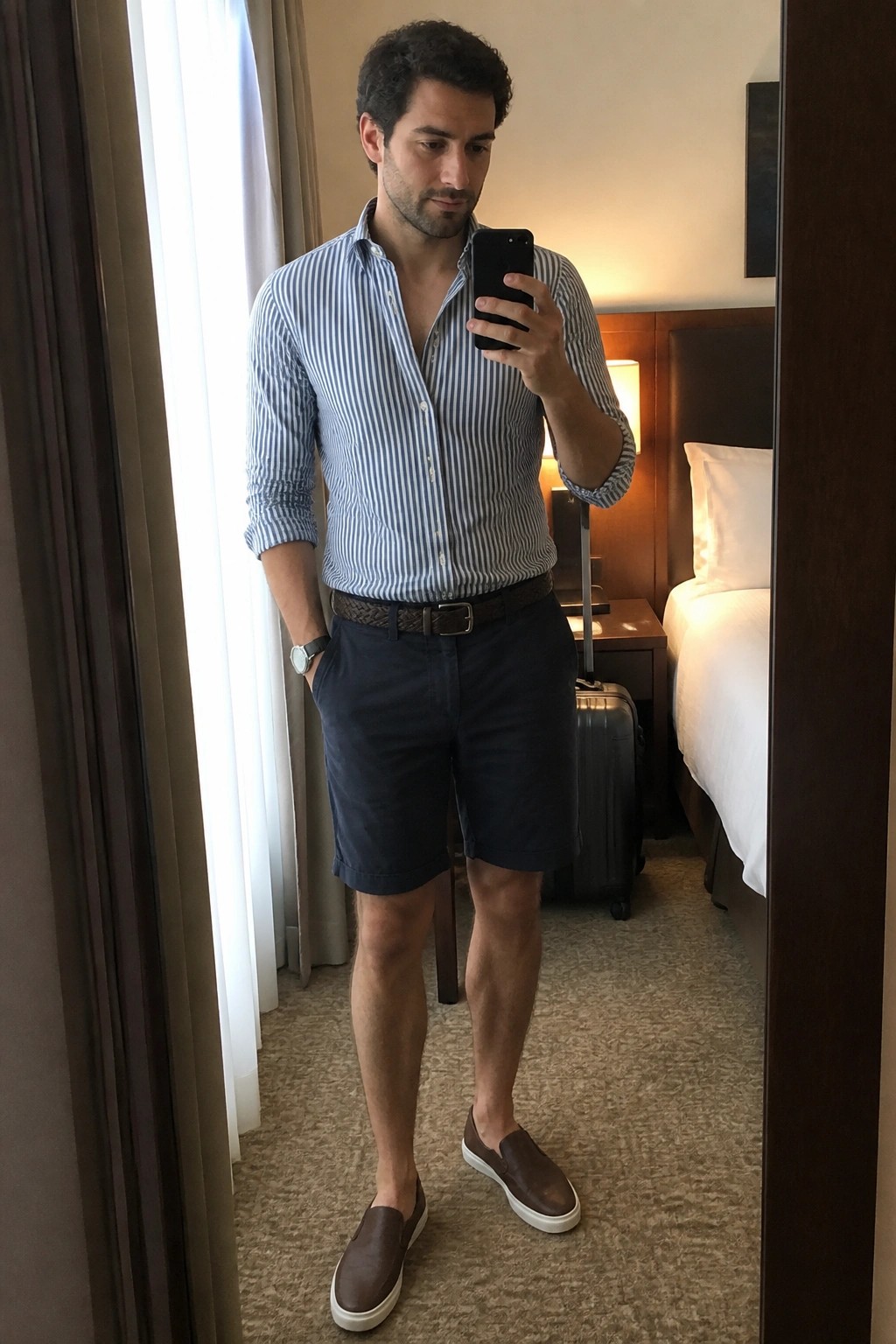 Man in striped shirt and shorts taking mirror selfie in hotel room